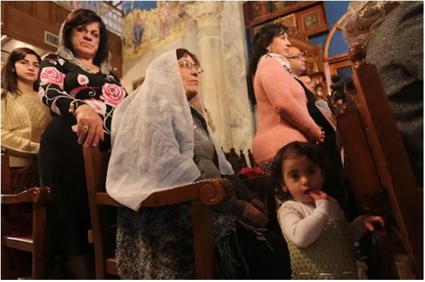 زنان، با سر پوشیده، همراه با کودکان در سکوهای کلیسا ایستاده اند.